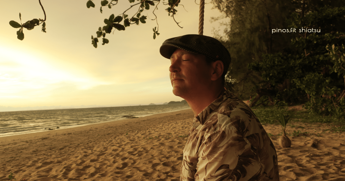 Mann sitzt mit geschlossenen Augen bei Sonnenuntergang am Strand und genießt ganz entspannt den Moment
